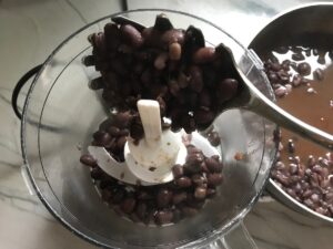 Spoon adding cooked black beans to food processor for refried black beans recipe.