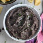 Creamy, rich, smokey, and delicious refried black beans recipe in a bowl with a few black beans and chopped cilantro sprinkled on top. On the counter are tortilla chips in a bowl.