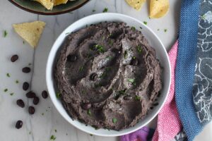 Creamy, rich, smokey, and delicious refried black beans recipe in a bowl with a few black beans and chopped cilantro sprinkled on top. On the counter are tortilla chips in a bowl.