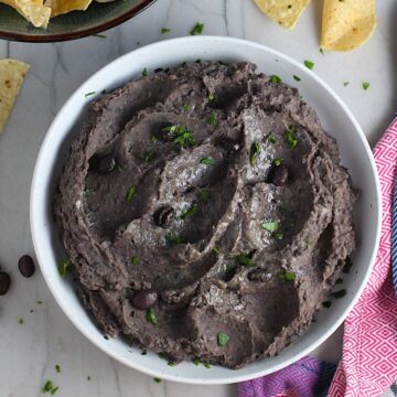 Creamy, rich, smokey, and delicious refried black beans recipe in a bowl with a few black beans and chopped cilantro sprinkled on top. On the counter are tortilla chips in a bowl.