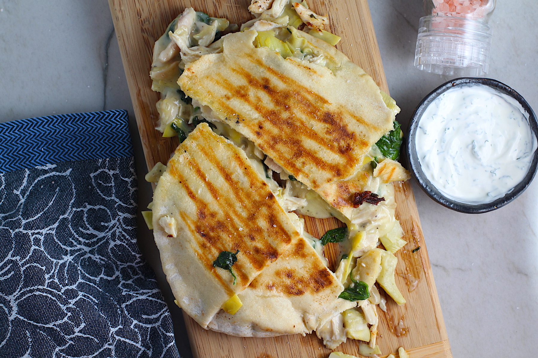 Spinach Artichoke Chicken Stuffed Pita is grilled and cut in half on a wood platter with filling oozing out. In the back is a small bowl of crema.
