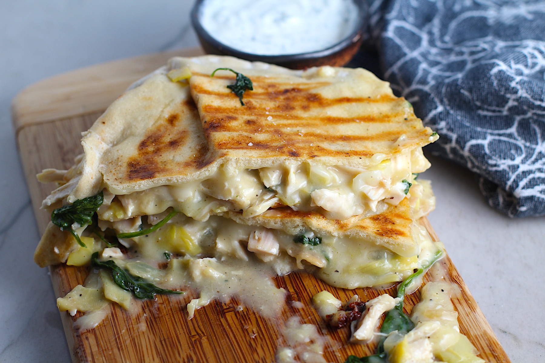 Spinach Artichoke Chicken Stuffed Pita is grilled and cut in half, then stacked on a wood platter with filling oozing out. In the back is a small bowl of crema.