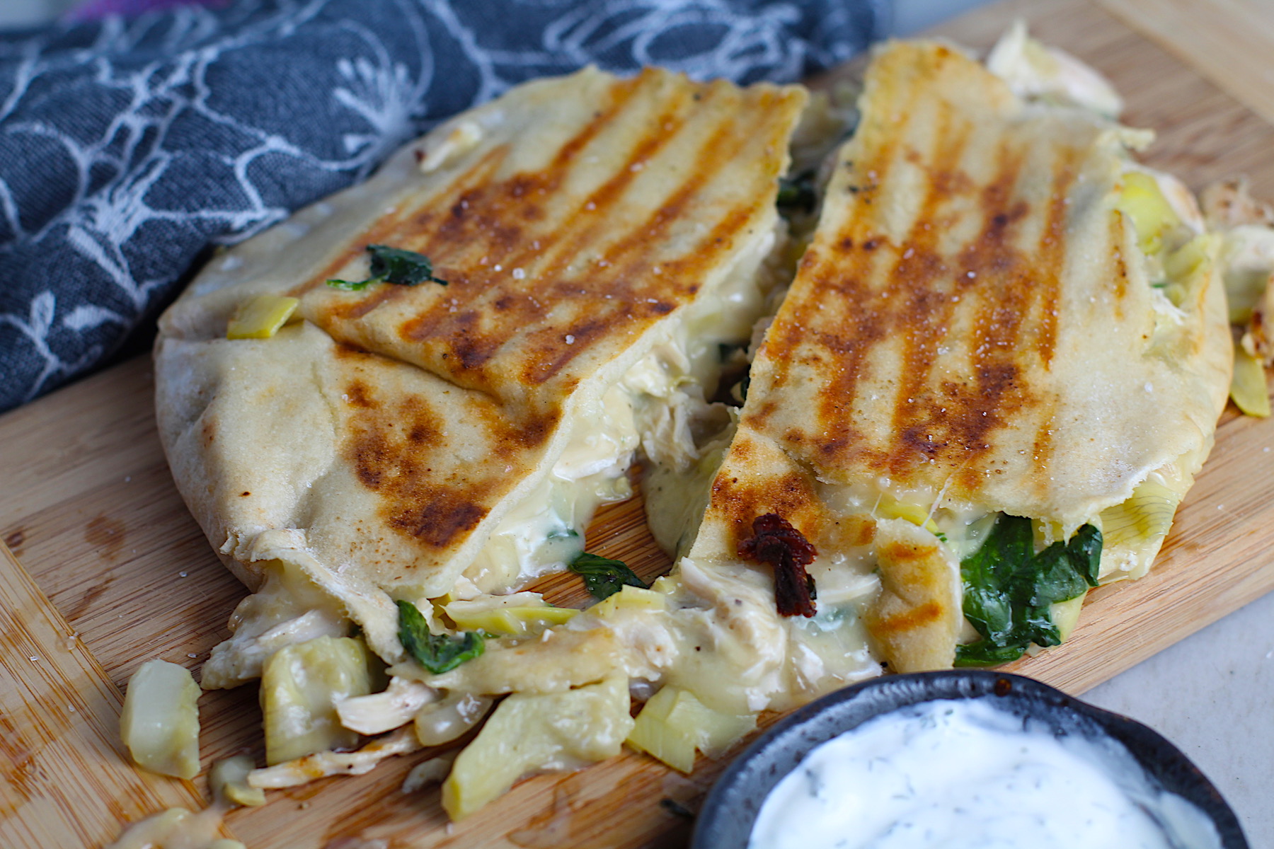 Spinach Artichoke Chicken Stuffed Pita is grilled and cut in half on a wood platter with filling oozing out. In the back is a small bowl of crema.