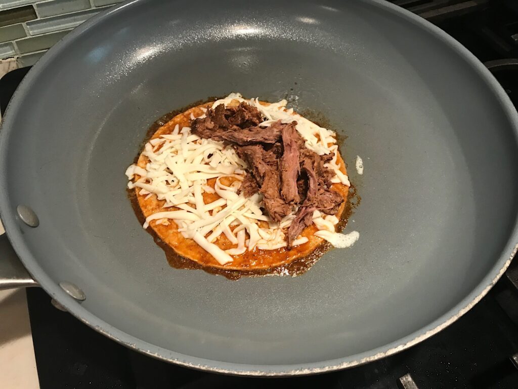 Corn tortilla with oaxaca cheese all over and shredded beef only on half in a hot pan for this Birria Tacos Recipe. Each taco has shredded beef and oaxaca cheese oozing out. On the plate is a bowl of red sauce, lime wedges, and cilantro.