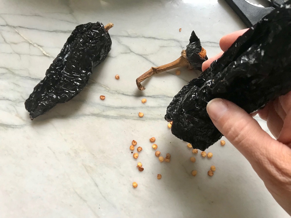 Hand shaking seeds out of dried ancho chile for Birria Tacos. This Birria Tacos Recipe in the Slow Cooker has shredded beef and Oaxaca cheese melted together in corn tortillas in a pan.