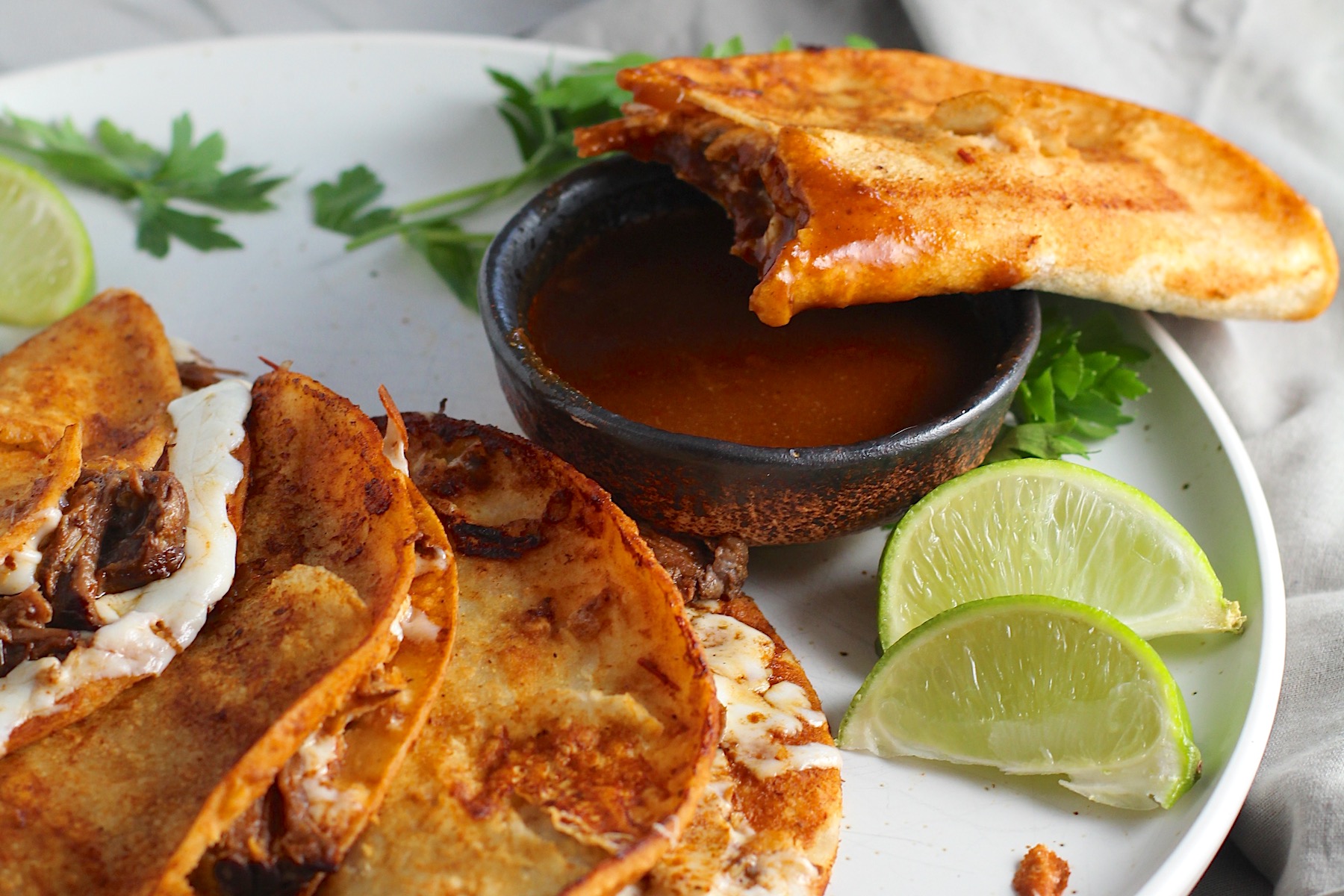 3 tacos fanned out on a plate with the 4th taco being dipped into sauce for this Birria Tacos Recipe. Each taco has shredded beef and oaxaca cheese oozing out. On the plate is a bowl of red sauce, lime wedges, and cilantro.