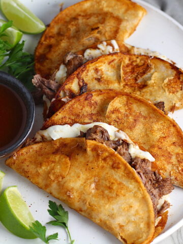 Four tacos fanned out on a plate for this Birria Tacos Recipe. Each taco has shredded beef and oaxaca cheese oozing out. On the plate is a bowl of red sauce, lime wedges, and cilantro.