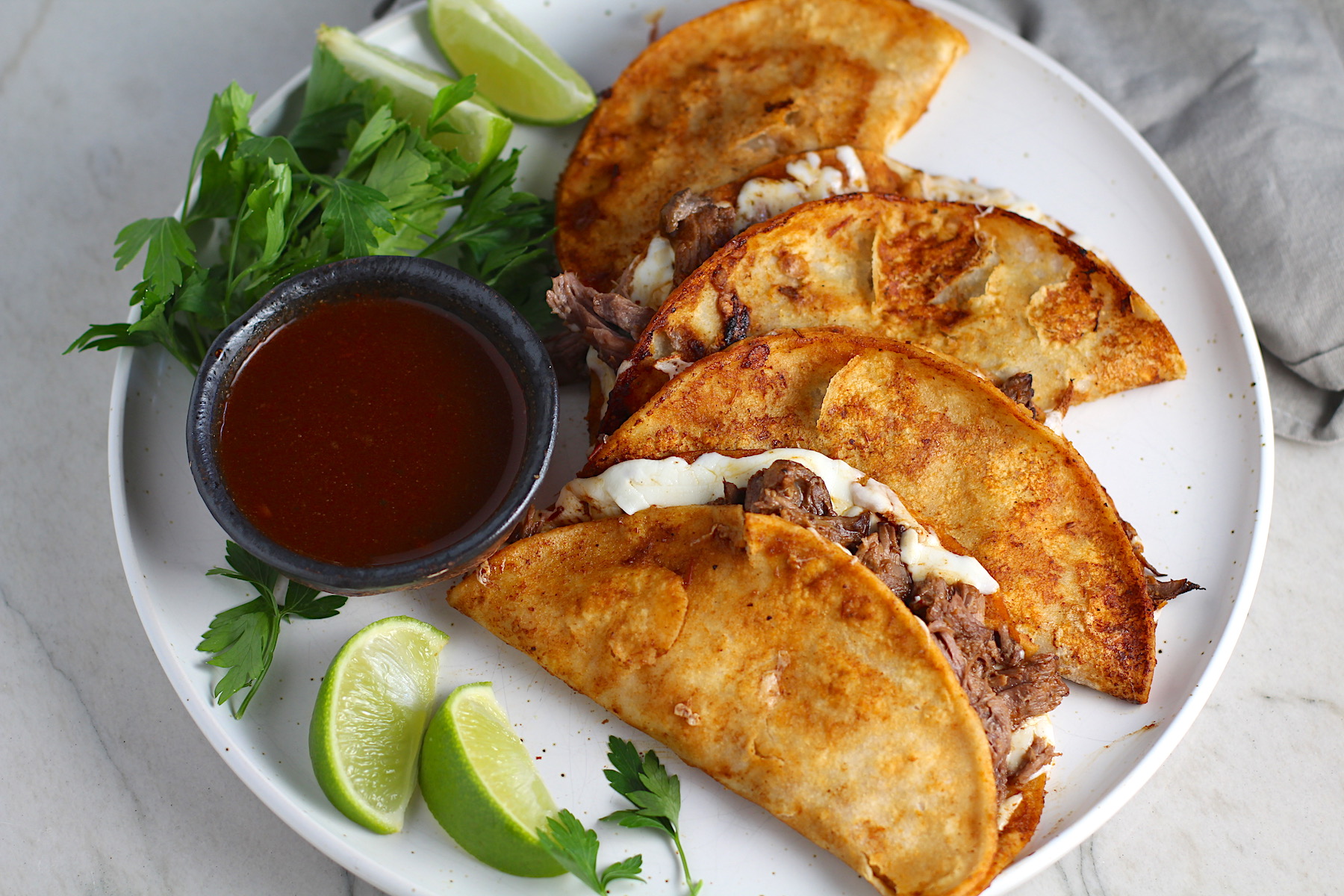 Four tacos fanned out on a plate for this Birria Tacos Recipe. Each taco has shredded beef and oaxaca cheese oozing out. On the plate is a bowl of red sauce, lime wedges, and cilantro.
