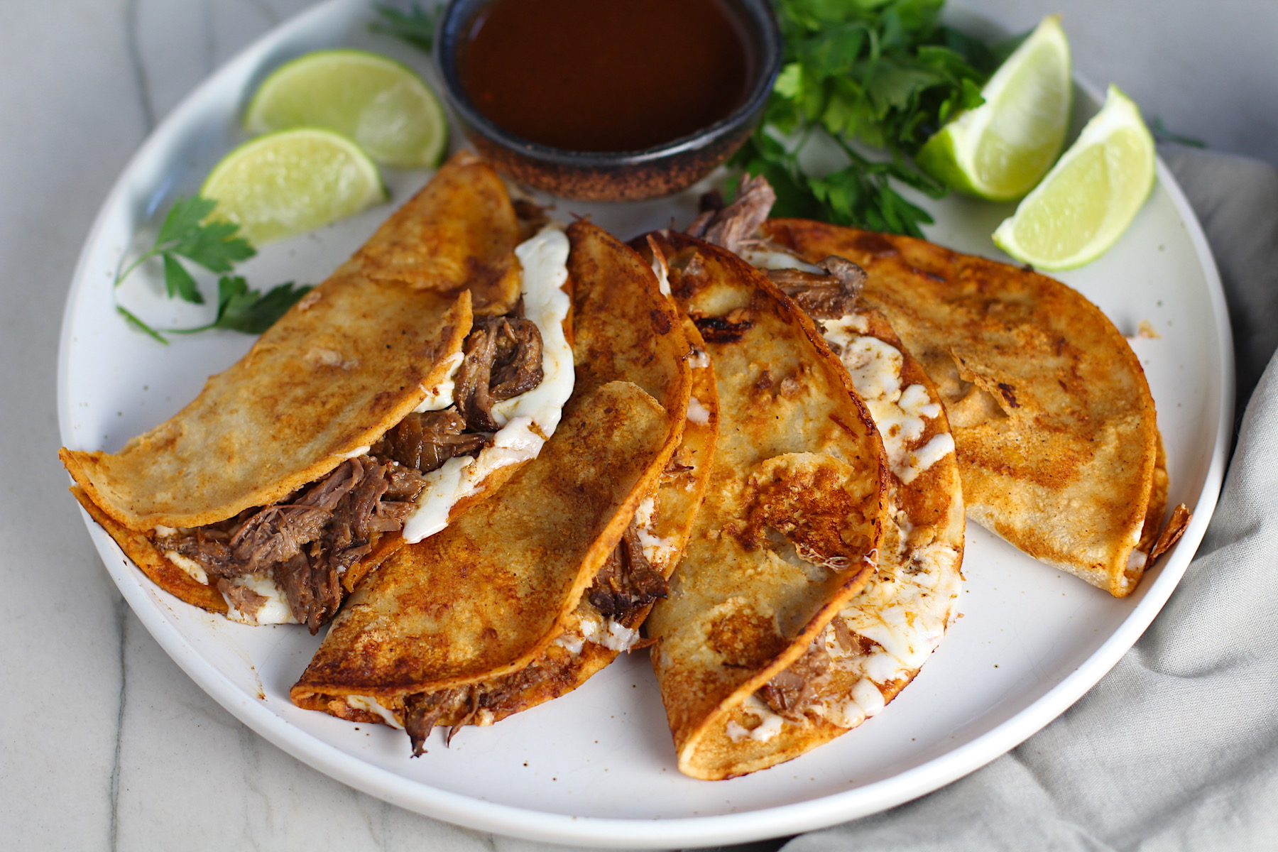 Four tacos fanned out on a plate for this Birria Tacos Recipe. Each taco has shredded beef and oaxaca cheese oozing out. On the plate is a bowl of red sauce, lime wedges, and cilantro.