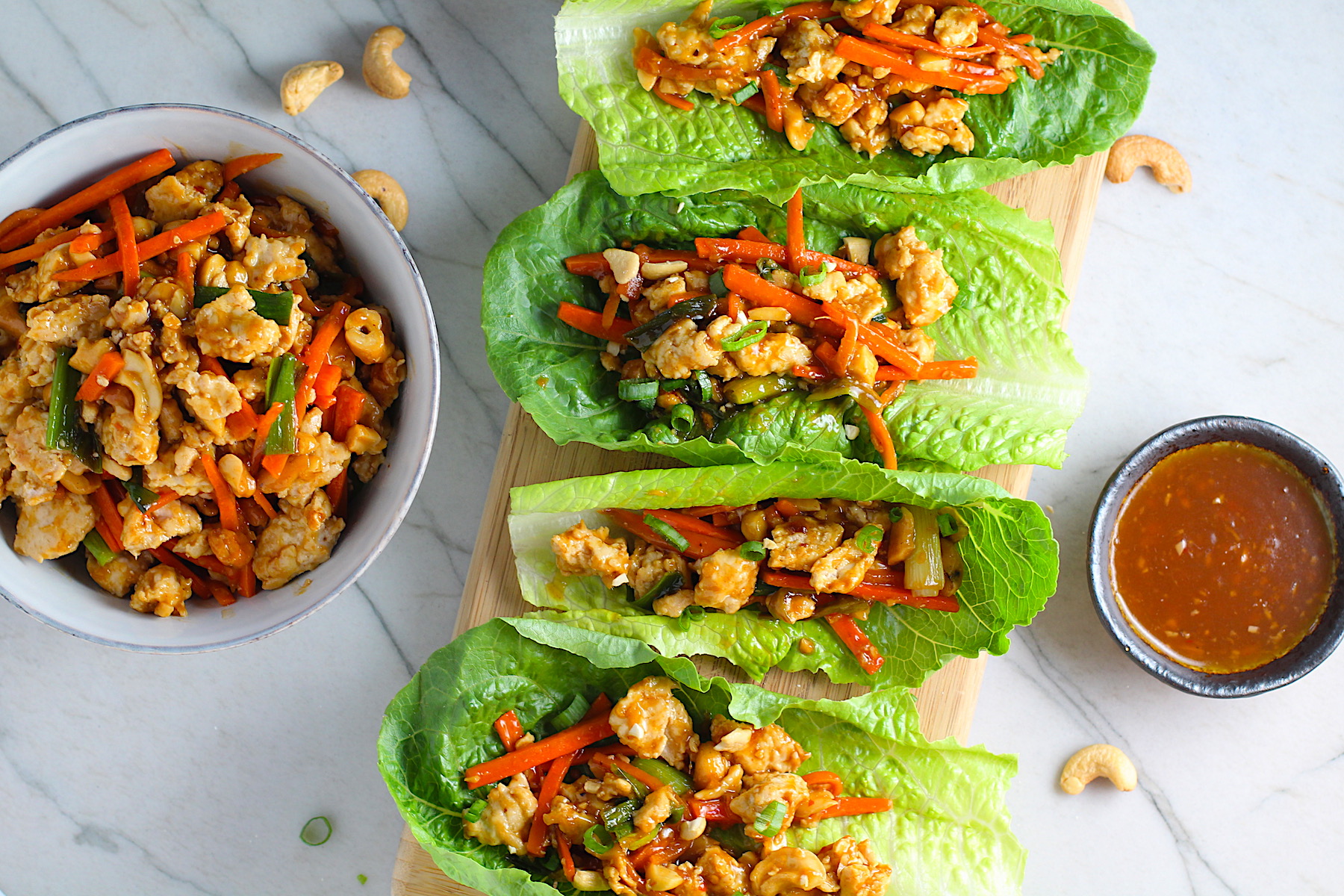 4 Low Carb Chicken Lettuce Wraps on counter with a bowl of Cashew Chicken and Carrot filling on left and sauce on right.