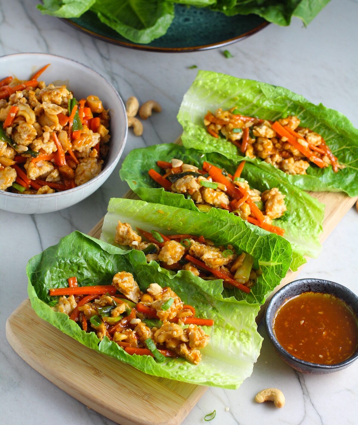 4 Low Carb Chicken Lettuce Wraps on counter with a bowl of Cashew Chicken and Carrot filling on left and sauce on right.