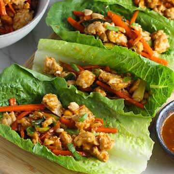 4 Cashew Low Carb Chicken Lettuce Wraps on counter with a bowl of Cashew Chicken and Carrot filling on left and sauce on right.