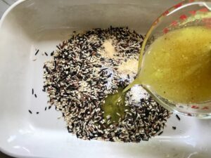 Measuring cup pouring broth into uncooked wild rice, garlic powder, and salt in a casserole dish for Chicken Wild Rice Casserole with Zucchini and Mushrooms.