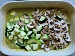 Uncooked triangle-cut zucchini and mushrooms added to casserole dish over rice and broth for Chicken Wild Rice Casserole with Zucchini and Mushrooms.