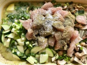 Raw cubed chicken with salt and pepper being added on top of zucchini and mushrooms added to casserole dish over rice and broth for Chicken Wild Rice Casserole with Zucchini and Mushrooms.