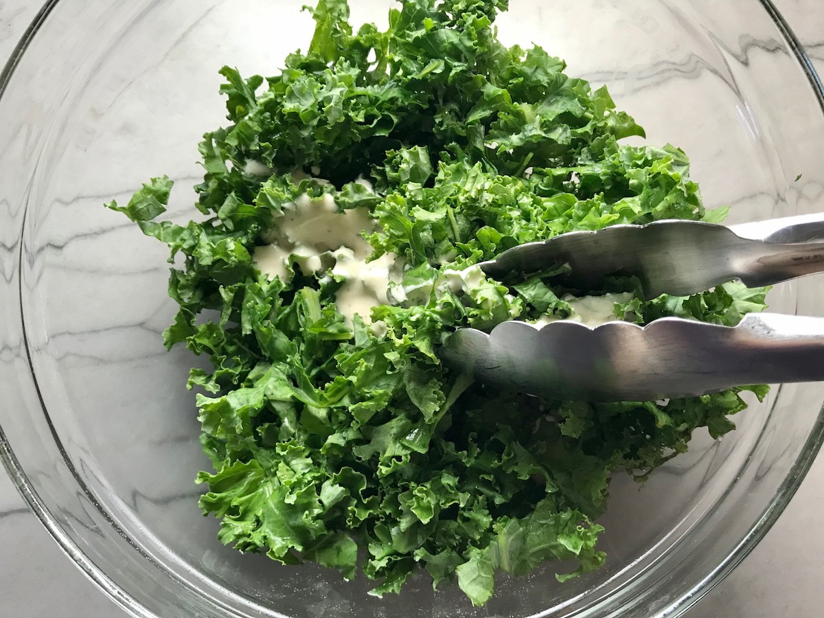 Tongs tossing kale with caesar dressing in a clear bowl for Kale Caesar Salad with Baked Crispy Lemon Chicken strips