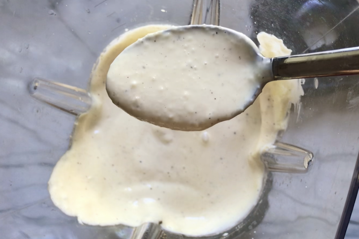 Blender with a spoon scooping blended Caesar dressing for Kale Caesar Salad with Baked Crispy Lemon Chicken strips