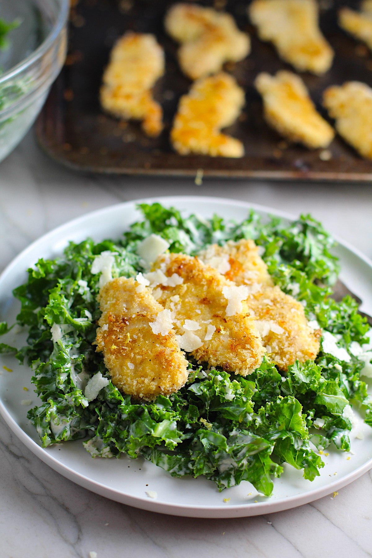 Crispy Lemon Chicken Kale Caesar Salad with chicken strips on top on a plate with sheet pan with chicken in the background.