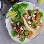Sheet Pan Pork Fajitas Recipe with red pepper, green pepper, and onion, in flour tortillas on a plate. Topped with Cilantro Lime Crema, fresh cilantro leaves, and lime wedges.