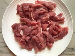 Strips of raw pork tenderloin on a plate for the Sheet Pan Pork Fajitas Recipe with red pepper, green pepper, and onion.