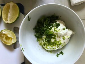Bowl with sour cream, cilantro, lime zest, and lime juice not mixed and lime cut in half next to bowl for the Sheet Pan Pork Fajitas Recipe with red pepper, green pepper, and onion.