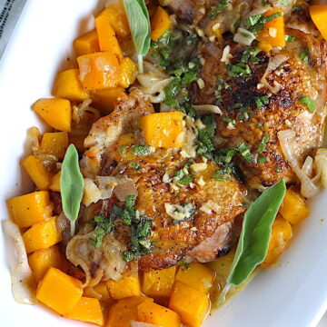Butternut squash and Chicken thighs with a sage, garlic, and shallot sauce on a platter with fresh sage leaves and topped with a buttery garlic, lemon, sage sauce. Lemon wedges, fresh sage leaves, and a kitchen towel on counter.