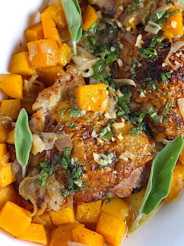 Butternut squash and Chicken thighs with a sage, garlic, and shallot sauce on a platter with fresh sage leaves and topped with a buttery garlic, lemon, sage sauce. Lemon wedges, fresh sage leaves, and a kitchen towel on counter.