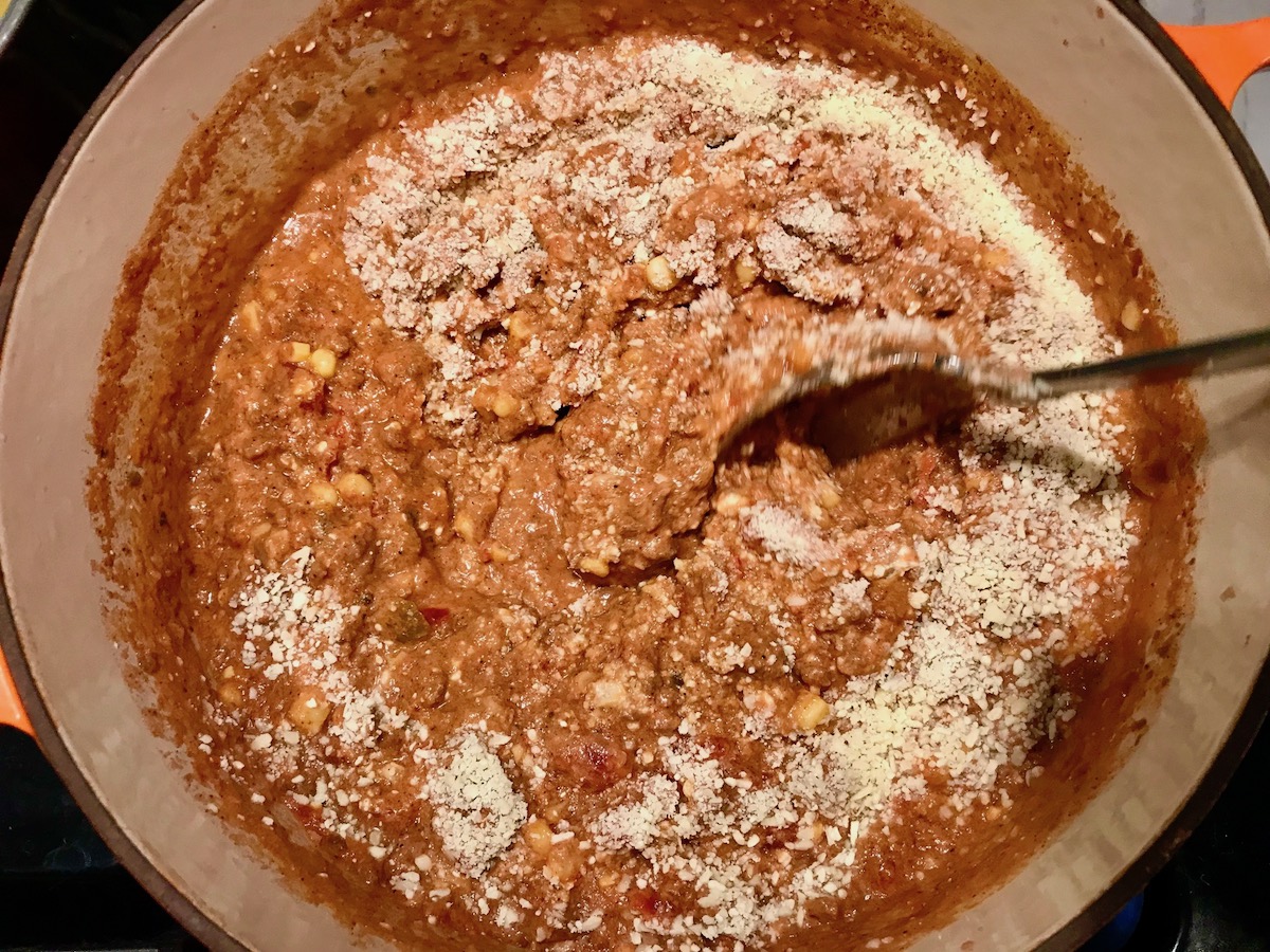 Spoon mixing parmesan cheese into pot of ground beef and tomato-chile sauce for Southwestern Pasta with Ground Beef and Corn and breadcrumbs. The sauce is thick and hearty with ground beef, sweet corn kernels, chunky tomatoes, chipotle peppers in adobo, green chiles, parmesan cheese, and lots of smokey and spicy seasonings.