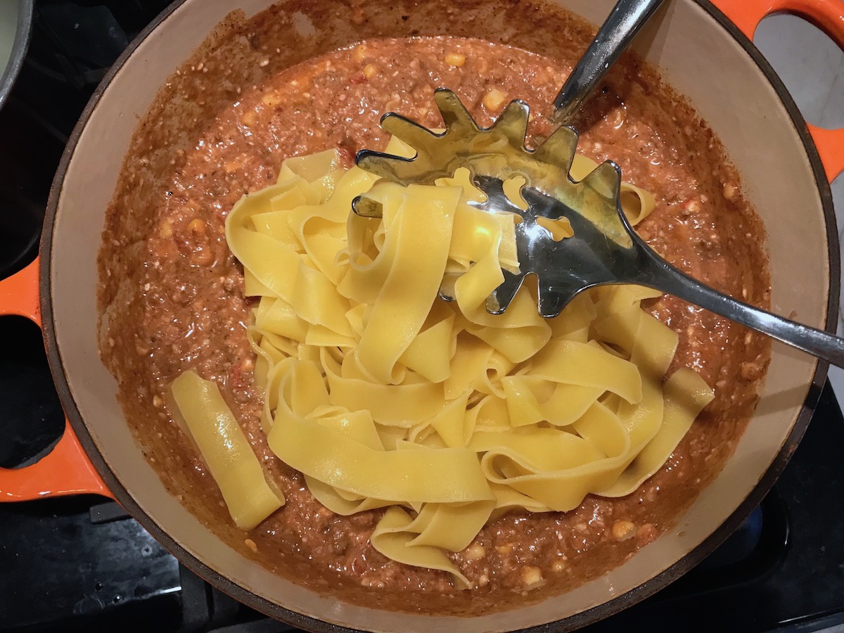 Pappardelle pasta being mixed into pot of ground beef and tomato-chile sauce for Southwestern Pasta with Ground Beef and Corn and breadcrumbs. The sauce is thick and hearty with ground beef, sweet corn kernels, chunky tomatoes, chipotle peppers in adobo, green chiles, parmesan cheese, and lots of smokey and spicy seasonings.