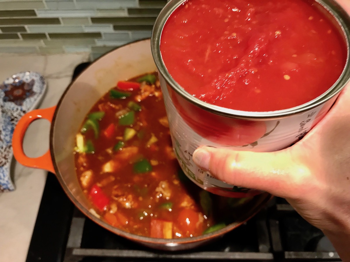 Hand adding can of diced tomatoes to pot on stove with red and green peppers, chicken, bacon and diced onions for Chicken Goulash with quick homemade dumplings.