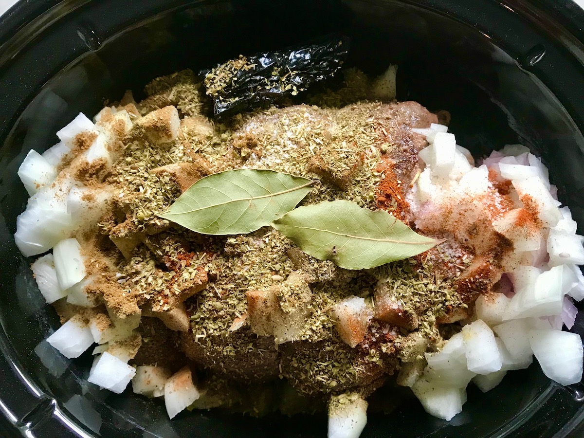 Beef roast, onion, seasonings, dried ancho chiles, and pay leaves in a slow cooker insert for Mexican Shredded Beef recipe.