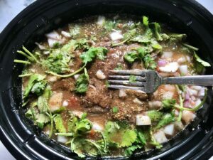 Fork pressing ingredients into liquid in slow cooker insert for Mexican Shredded Beef recipe.