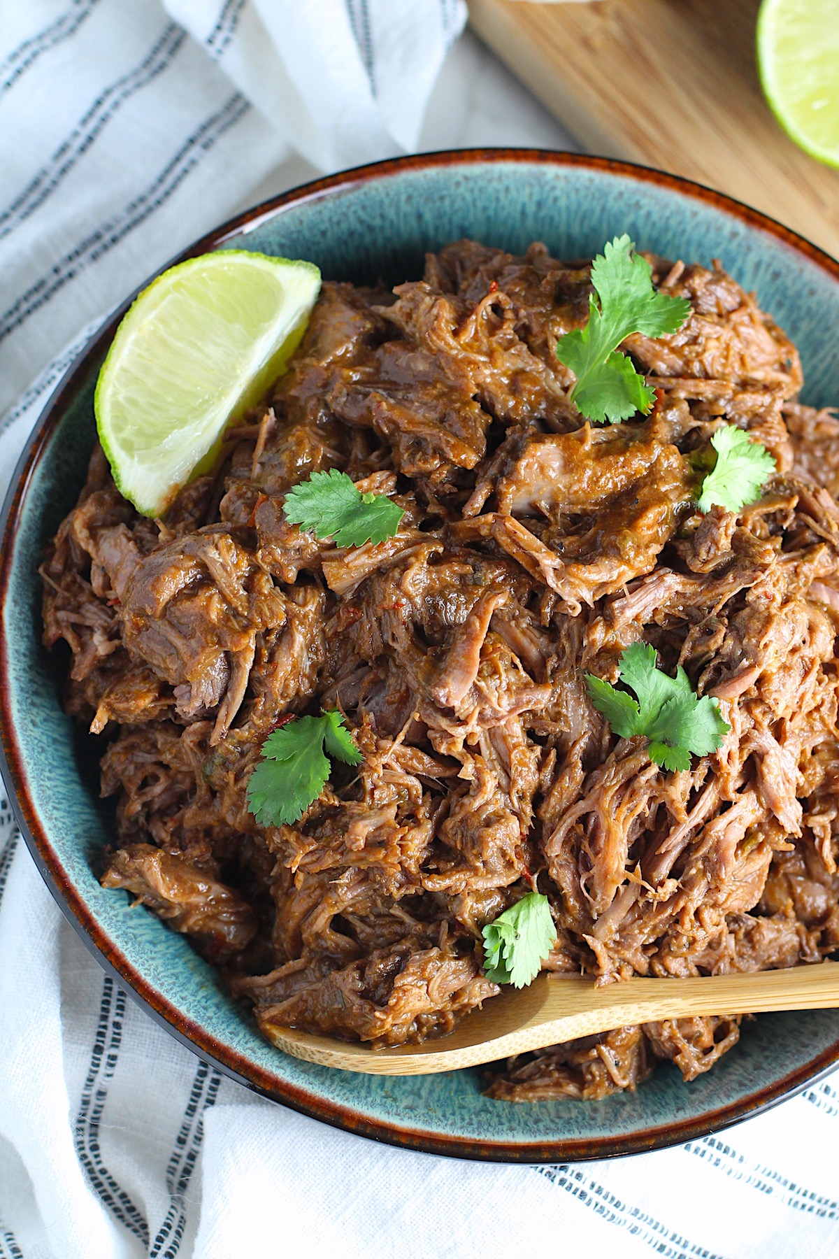 Mexican Shredded Beef in a serving bowl with cilantro on top, lime wedge on side and wood serving spoon in bowl. it's incredibly easy to make in a slow cooker or in the oven and can be used for quick and easy family weeknight dinners! 