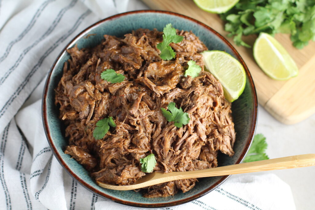 Mexican Shredded Beef in a serving bowl with cilantro on top, lime wedge on side and wood serving spoon in bowl. it's incredibly easy to make in a slow cooker or in the oven and can be used for quick and easy family weeknight dinners!
