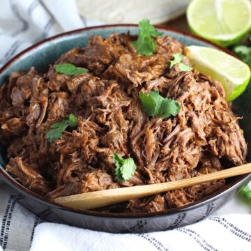 Mexican Shredded Beef in a serving bowl with cilantro on top, lime wedge on side and wood serving spoon in bowl. it's incredibly easy to make in a slow cooker or in the oven and can be used for quick and easy family weeknight dinners! 