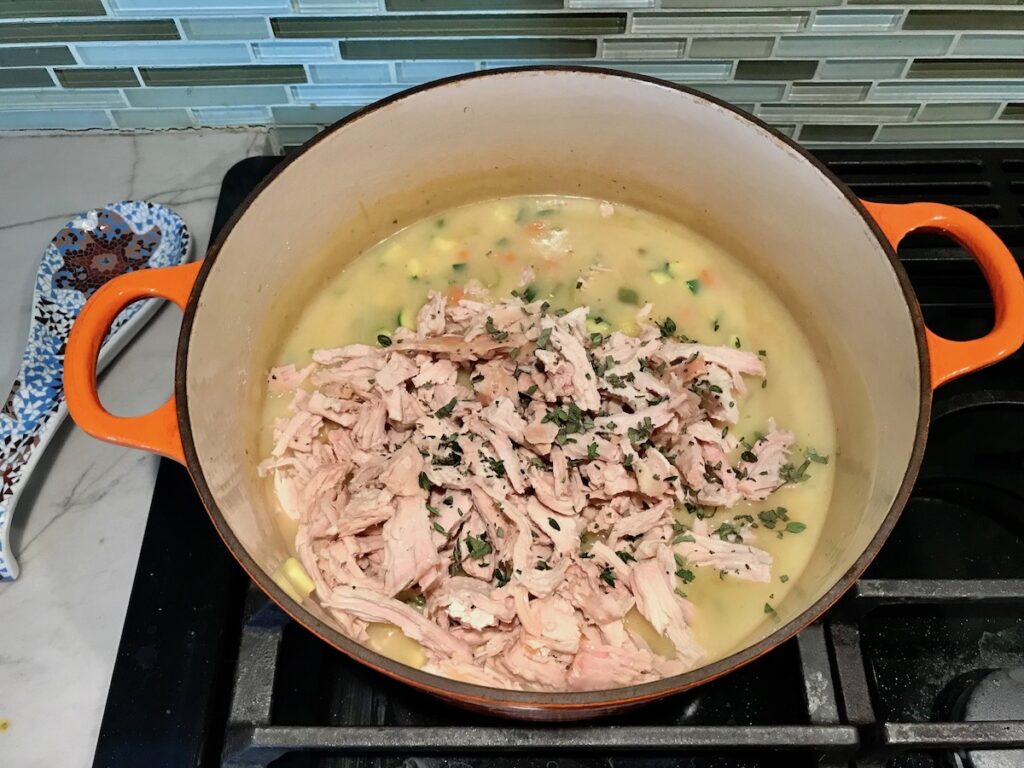 Shredded turkey and herbs added on top of sauce in pot for Puff Pastry Turkey Pot Pie. It's an easy dinner recipe to use leftover turkey from the holidays and ingredients you can have on hand! The vegetables and sauce come together so easily and store-bought puff pastry is a time saver.