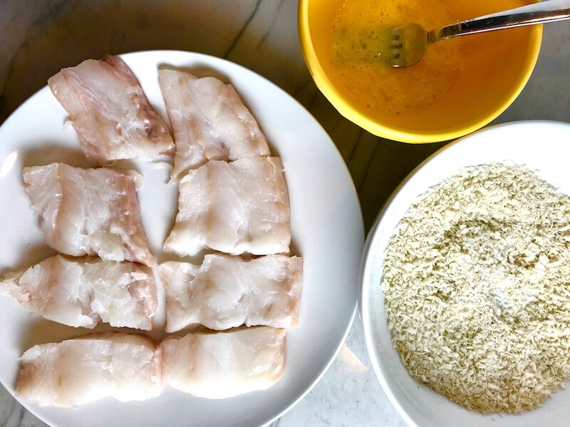 Raw cod fish cut into large pieces on a plate next to egg and breading stations for Crispy Baked Cod Panko Fish and Chips.