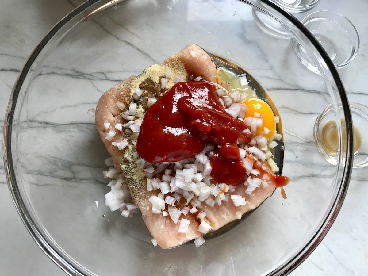 Raw ground chicken, diced onion, egg, and ketchup in a bowl for Ground Chicken Meatloaf. It's a perfect family dinner main dish! 10 minutes to prep and 45 minutes to bake.