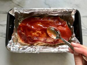 Spoon spreading ketchup over the top of raw Ground Chicken Meatloaf in a loaf pan that's lined with aluminum foil. It's a perfect family dinner main dish! 10 minutes to prep and 45 minutes to bake.