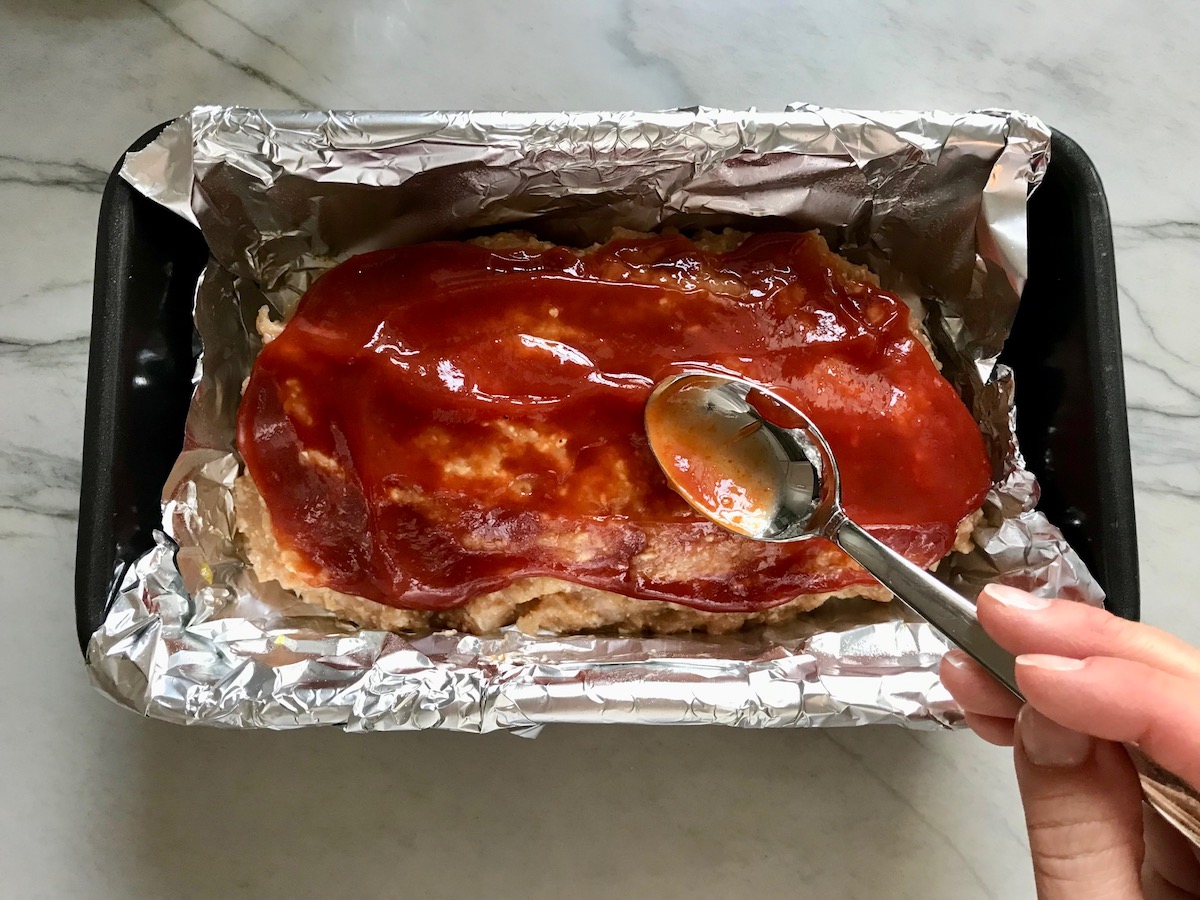 Spoon spreading ketchup over the top of raw Ground Chicken Meatloaf in a loaf pan that's lined with aluminum foil. It's a perfect family dinner main dish! 10 minutes to prep and 45 minutes to bake.