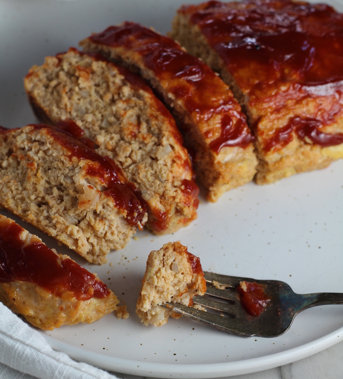 Ground Chicken Meatloaf with ketchup on top sliced and fanned out on plate with fork and a piece on side and towel next plate. It's a perfect family dinner main dish! 10 minutes to prep and 45 minutes to bake.