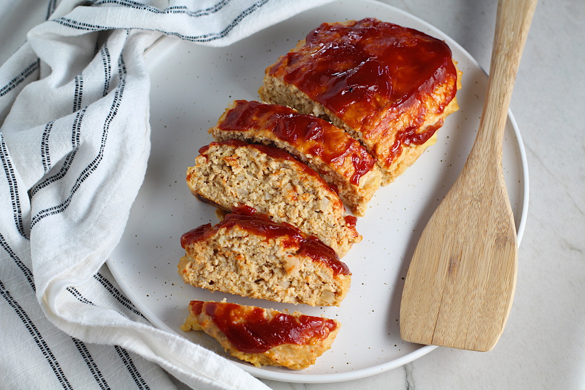 Ground Chicken Meatloaf with ketchup on top sliced and fanned out on plate with spatula on side and towel next plate. It's a perfect family dinner main dish! 10 minutes to prep and 45 minutes to bake.