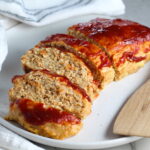 Ground Chicken Meatloaf with ketchup on top sliced and fanned out on plate with spatula on side and towel next plate. It's a perfect family dinner main dish!  10 minutes to prep and 45 minutes to bake.