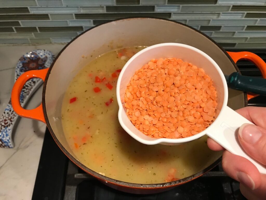 Hand holding measuring cup of dry red lentils over pot of soup for Red Pepper and Lentil Soup. This is an easy and hearty lunch or dinner! It comes together in just 40 minutes.