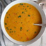 Creamy Red Pepper and Lentil Soup in a bowl with spoon and pot in background. Whole red pepper next to bowl. This is an easy and hearty lunch or dinner!  It comes together in just 40 minutes.