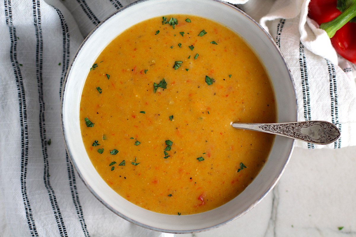 Creamy Red Pepper and Lentil Soup in a bowl with spoon and pot in background. Whole red pepper next to bowl. This is an easy and hearty lunch or dinner! It comes together in just 40 minutes.