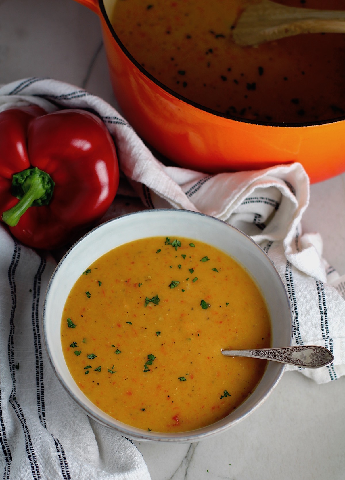 Creamy Red Pepper and Lentil Soup in a bowl with spoon and pot in background. Whole red pepper next to bowl. This is an easy and hearty lunch or dinner! It comes together in just 40 minutes.
