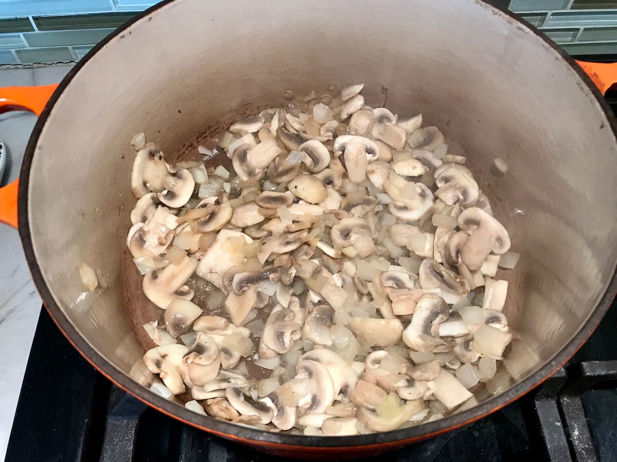 Sliced mushrooms and diced onions cooking in a pot on stove for Chicken Stroganoff Brazilian Style. It's an easy 30 minute dinner with a tomato based sauce, cream, and mushrooms all over rice.