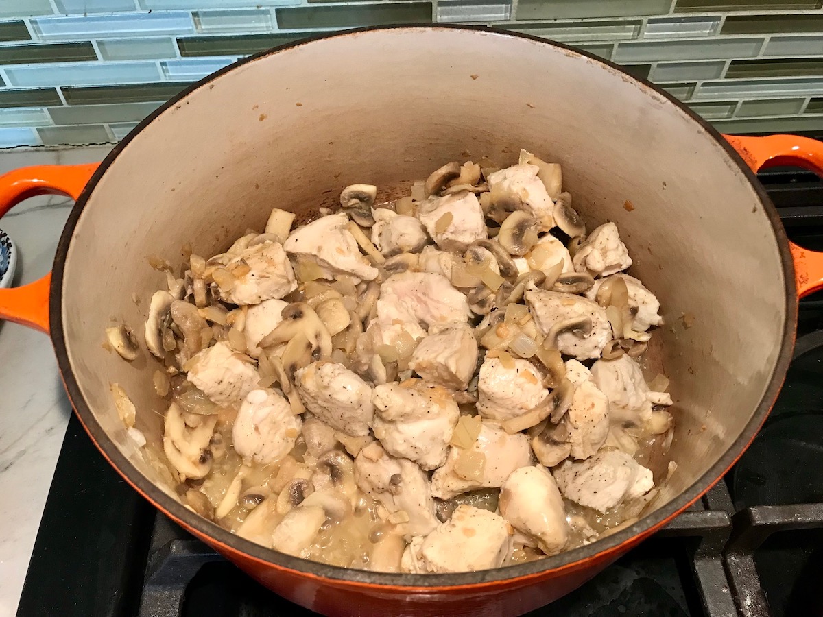 Chicken chunks, sliced mushrooms and diced onions cooking in a pot on stove for Chicken Stroganoff Brazilian Style. It's an easy 30 minute dinner with a tomato based sauce, cream, and mushrooms all over rice.