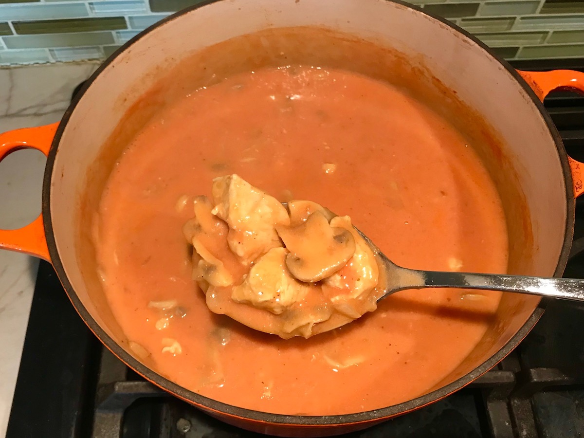 Spoon with scoop of chicken and mushroom over a pot on stove for Chicken Stroganoff Brazilian Style. It's an easy 30 minute dinner with a tomato based sauce, cream, and mushrooms all over rice.